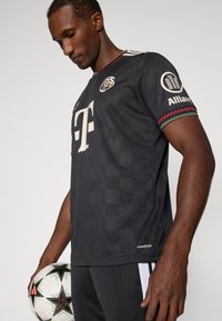 Camiseta deportiva negra de manga corta con un gran logo beige, hombros acentuados y ribetes rojos con patrón. El modelo sostiene un balón de fútbol blanco.