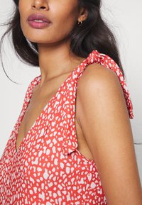 Rode stoffen jurk met witte abstracte stippen, met strikbanden op de schouders en een diepe V-halslijn. Soepele textuur overal.