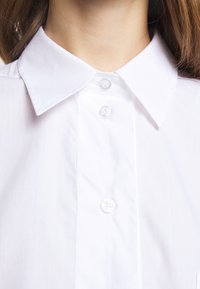 Primer plano de una persona que lleva una camisa blanca de cuello con botones, mostrando tres botones visibles y parte del cuello y el cabello.