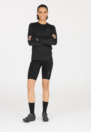 Schwarzes, sportliches Langarmoberteil und enganliegende Shorts, beide aus einem glatten Stoff. Außerdem schwarze Radschuhe und dunkle Socken.