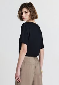 Donna con capelli corti castani, che indossa una maglietta nera a maniche corte e pantaloni beige a vita alta, che guarda sopra la spalla contro uno sfondo semplice.