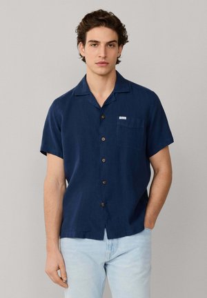 Joven con cabello rizado oscuro, vestido con una camisa azul marino de manga corta con botones y jeans azul claro, de pie con una mano en el bolsillo.