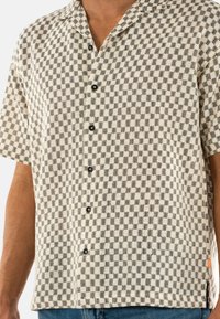 Camicia a maniche corte con colletto a bottoni, caratterizzata da un motivo a quadretti bianco e nero, realizzata in tessuto testurizzato, con bottoni neri e un'etichetta arancione.