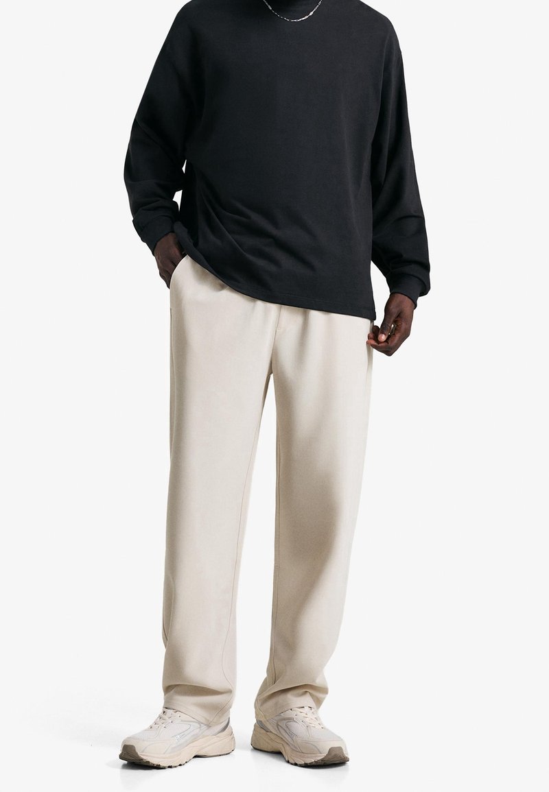 Personne portant une chemise noire à manches longues, un pantalon beige clair et des baskets beiges, debout avec les mains dans les poches sur un fond blanc uni.