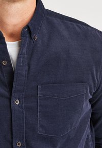 Marineblauwe corduroy button-up shirt met een borstzak, contrasterende stiksels en een kraag. Zachte textuur met subtiele ribbels.