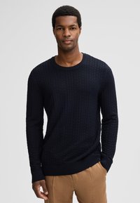 Mężczyzna ubrany w granatowy, textured sweter z długim rękawem i jasnobrązowe spodnie, stojący z jedną ręką w kieszeni na jednolitym tle.