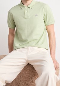 Polo en coton vert clair avec un col classique et trois boutons. Présente un petit logo brodé sur la poitrine. Porté avec un pantalon de couleur crème.