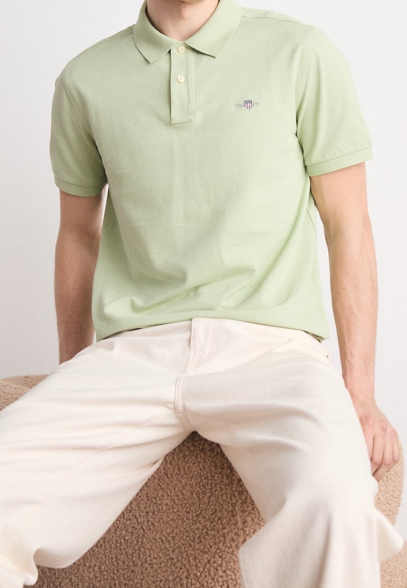 Polo en coton vert clair avec un col classique et trois boutons. Présente un petit logo brodé sur la poitrine. Porté avec un pantalon de couleur crème.