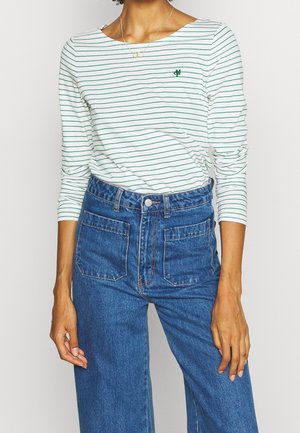 Femme portant une chemise blanche à manches longues avec de fines rayures vertes et un logo vert brodé, associée à un jean bleu taille haute avec des poches plaquées à l'avant.