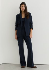 Donna con capelli lunghi e scuri che indossa una giacca blu navy, pantaloni larghi coordinati e scarpe marroni, in piedi contro un muro bianco.