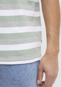Camiseta de algodón con rayas horizontales en verde claro, gris y blanco. Presenta un dobladillo recto y una etiqueta en el costado. Se lleva con jeans de mezclilla azul.