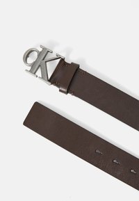 Ceinture en cuir marron avec une boucle en métal argenté en forme de lettre "K", ornée de coutures discrètes le long des bords et de trous ajustables.