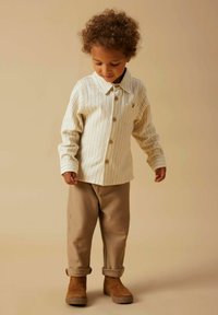 Camicia a strisce chiare con taschino sul petto, abbinata a pantaloni beige con risvolto e stivali alla caviglia marroni, su uno sfondo uniforme.