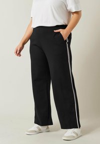 Pantaloni neri a gamba larga con strisce bianche sui lati, dotati di tasche laterali. Realizzati in materiale morbido, con una vestibilità comoda. Indossati con sneakers bianche.