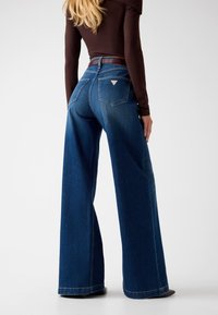 Denim wijde pijpen jeans in donkerblauw met een hoge taille, voorzien van een bruine riem en achterzakken. Gladde textuur en losse silhouet.