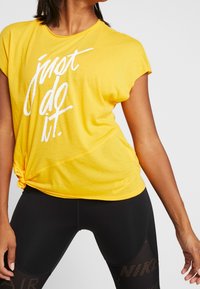 Camiseta amarilla de manga corta con el texto impreso en blanco "just do it" y un detalle anudado en la cintura, combinada con leggins negros.