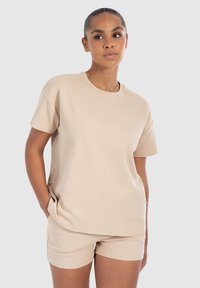 Beige lyhythihainen t-paita, joka on valmistettu pehmeästä, tekstuurista kankaasta. Siinä on pyöreä pääntie ja hienovarainen brodeerattu logo rinnassa.