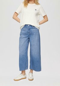 Valkoinen puuvillainen t-paita, jossa pieni sydänlogo, yhdistetty vaaleansinisiiin leveälahkeisiin farkkuihin ja beigen värisiin tennareihin, joissa on teksturoidut yksityiskohdat.