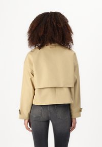 Veste cropped kaki avec un design à dos superposé, manches longues et poignets boutonnés, associée à un jean ajusté gris foncé, texture lisse.