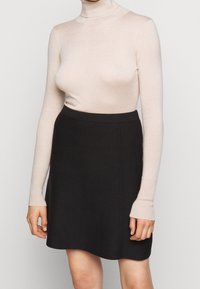 Kvinna som bär en beige långärmad polotröja och en högmidjad svart kjol står mot en enkel bakgrund.