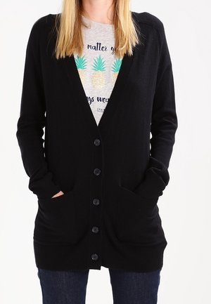 Femme portant un cardigan noir boutonné sur une chemise grise avec trois ananas et du texte, mains dans les poches du cardigan, jean bleu.