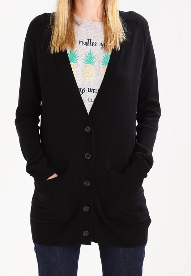 Femme portant un cardigan noir boutonné sur une chemise grise avec trois ananas et du texte, mains dans les poches du cardigan, jean bleu.