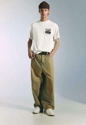 Biała koszulka z czarnym nadrukiem Forda, zestawiona z oversize'owymi jasnobrązowymi spodniami i białymi sneakersami. Brązowa czapka w paski.