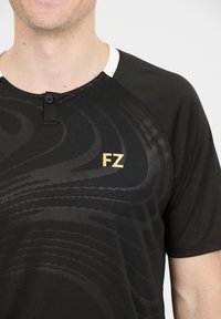 Musta lyhythihainen paita, jossa on sävytetty aalto-kuvio, pyöreä pääntie ja kaksinappinen lista. Edessä on kultainen "FZ"-logo.