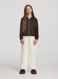 Giacca marrone con zip e cappuccio in tessuto strutturato, abbinata a pantaloni bianchi a gamba larga e scarpe slip-on marroni. Sfondo neutro.