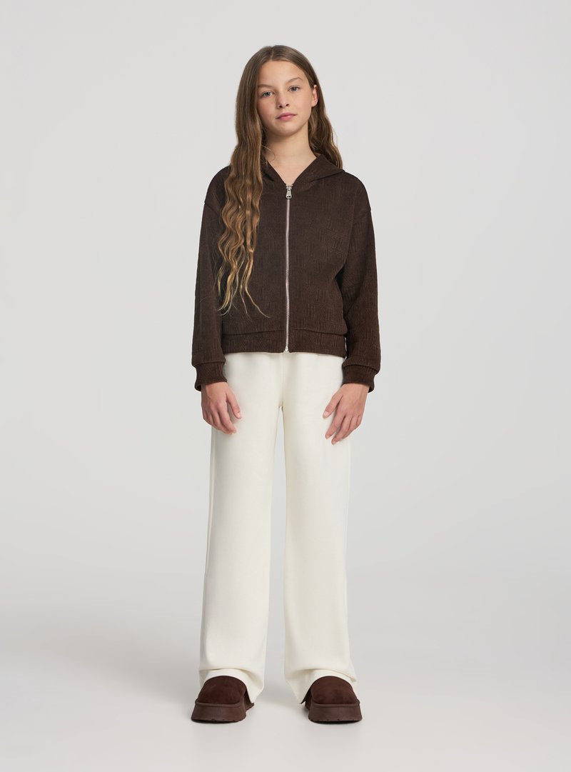 Giacca marrone con zip e cappuccio in tessuto strutturato, abbinata a pantaloni bianchi a gamba larga e scarpe slip-on marroni. Sfondo neutro.