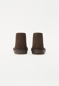 Brūni zamšādas potītes zābaki ar plakanu zoli, kuriem ir tīrs dizains un minimālas šuves gar malām. Nav pamanāmu aparatūras vai akcentu.