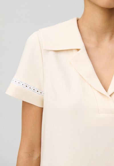 Blusa color crema con cuello, mangas cortas con detalles de encaje blanco y un tejido texturizado. Diseño frontal sin botones.