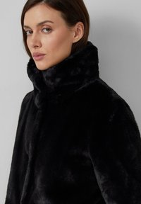 Manteau en fausse fourrure noire avec un col haut, texture lisse et design épuré. Dispose d'une fermeture à l'avant et sans détails matériels visibles.