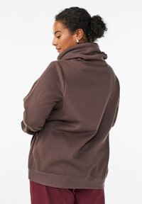 Sudadera con capucha de gran tamaño en color marrón, con un alto cuello, textura suave y dobladillo acanalado, combinada con pantalones de color marrón oscuro. Sin patrones ni acentos visibles.