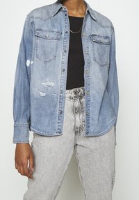 Jasnoniebieska koszula dżinsowa o wzornictwie inspirowanym stylem western, z dwiema kieszeniami na piersi, widocznym przeszyciem i przetarciami z przodu.