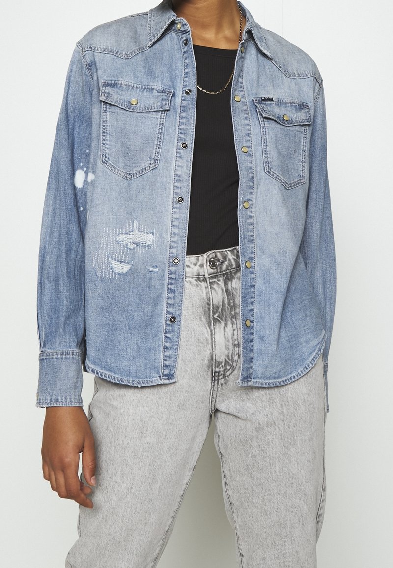 Jasnoniebieska koszula dżinsowa o wzornictwie inspirowanym stylem western, z dwiema kieszeniami na piersi, widocznym przeszyciem i przetarciami z przodu.