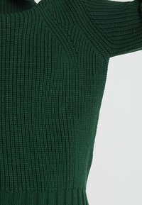 Maglione lavorato a maglia verde scuro con texture a coste, caratterizzato da polsini aderenti e un collo alto. Dettagli delle cuciture visibili lungo le spalle.