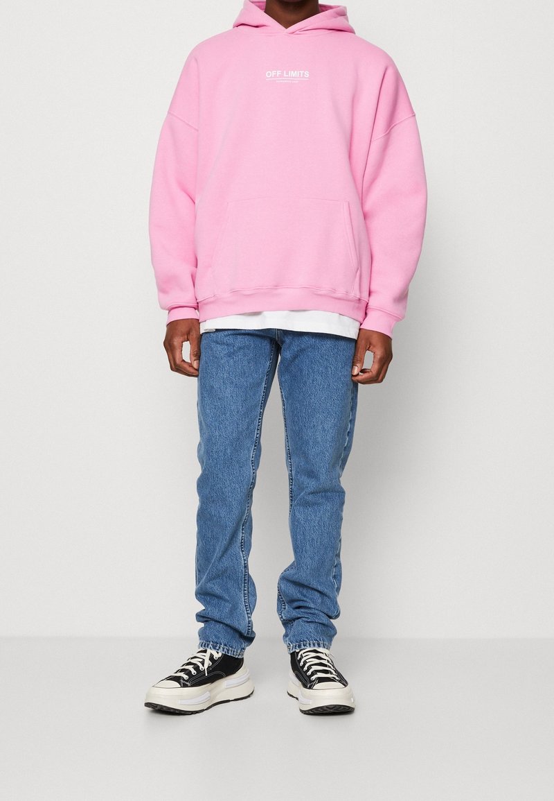 Personne portant un sweat à capuche rose avec l'inscription "OFF LIMITS", un jean bleu et des baskets noir et blanc, debout devant un fond blanc uni.