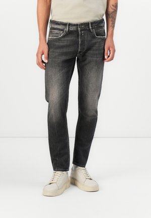 Mies pukeutunut haalistuneisiin mustiin suoralahkeisiin farkkuihin ja valkoisiin lenkkareihin, seisoo yksinkertaisen valkoisen taustan edessä.