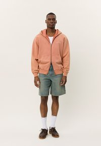 Breskvas zip-up hoodie s prednjim džepom, nosi se preko bijelog T-shirt-a, uparen s trapericama i smeškim cipelama, postavljen na neutralnoj pozadini.
