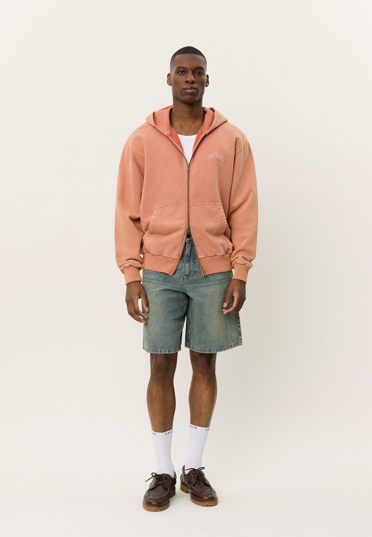 Perzikkleurige zip-up hoodie met een voorzak, gedragen over een wit T-shirt, gecombineerd met spijkerbroek-shorts en bruine schoenen, scherp afgetekend tegen een neutrale achtergrond.