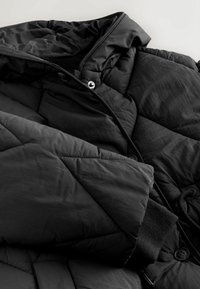 Mustast polsterdatud jakk, millel on teemanttikandiga tekstuur, eemaldatav kapuuts, pressnööbid ja elastsed varrukapitsid mugavaks istuvuseks.