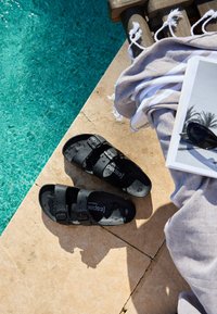 Black wet sandals on poolside stone tiles next to a light gray towel and a book with sunglasses resting on it.