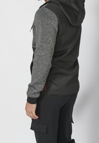 Homme portant un sweat à capuche noir avec des manches grises et un pantalon cargo noir, debout de profil avec les mains détendues le long du corps.