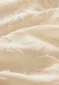 Tissu couleur crème avec des motifs floraux en relief et des détails de broderie texturés et duveteux sur un fond doux et lisse.