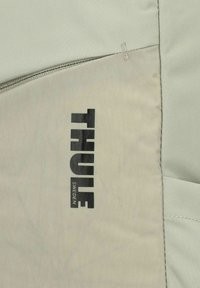 Sac à dos Thule en tissu gris clair, présentant un design texturé et un logo noir proéminent avec "SUÈDE" en dessous, fermé par une fermeture éclair.