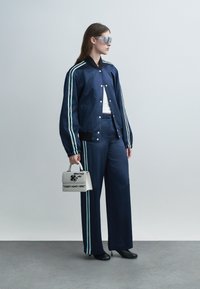 Kobieta ubrana w granatową kurtkę i spodnie z jasnoniebieskimi paskami, trzymająca białą torebkę z napisem oraz nosząca duże, refleksyjne okulary przeciwsłoneczne.