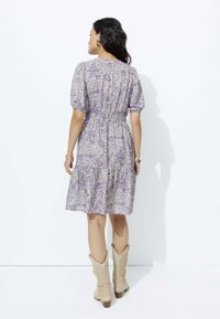 Robe florale avec un motif violet et crème, manches courtes à bouffant, taille cintrée, jupe à volants, portée avec des bottes de cowboy beiges.