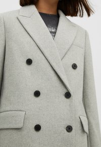 Vrouw met een lichtgrijze wollen mantel met dubbele rij knopen, zwarte knopen en klepzakken, gedragen over een zwarte top met pailletten.