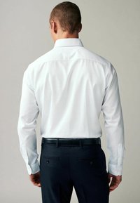 Chemise blanche à manches longues avec boutons, arborant un col classique et une texture lisse, associée à un pantalon sombre et une ceinture noire.